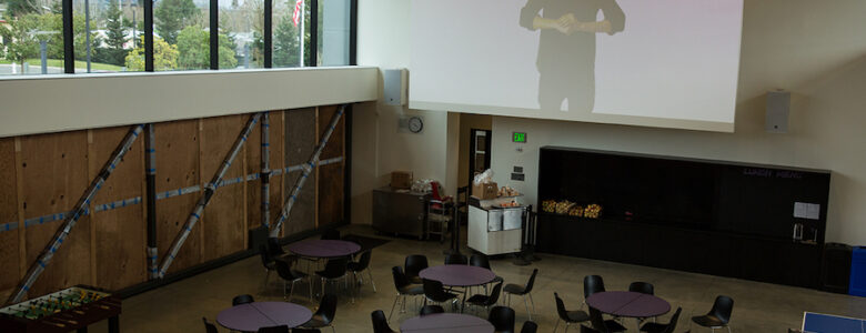 gathering space with several round tables and students looking at a large projection screen